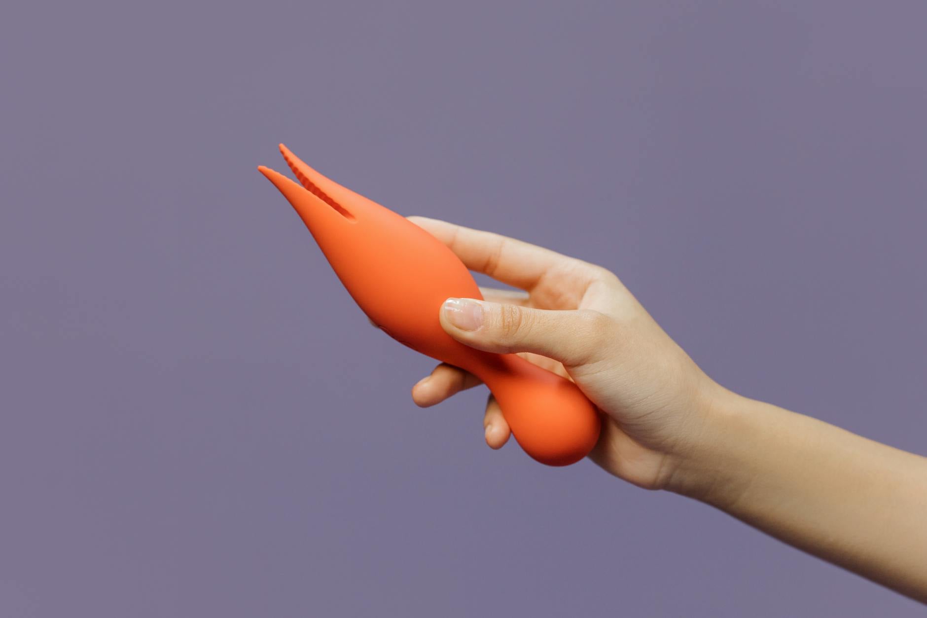 A hand holding a vibrant clitoral vibrator against a minimalistic purple backdrop, showcasing personal intimacy.